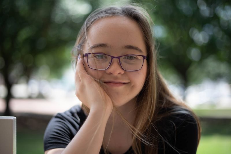 A smiling young woman with Down syndrome rests her chin on her hand.