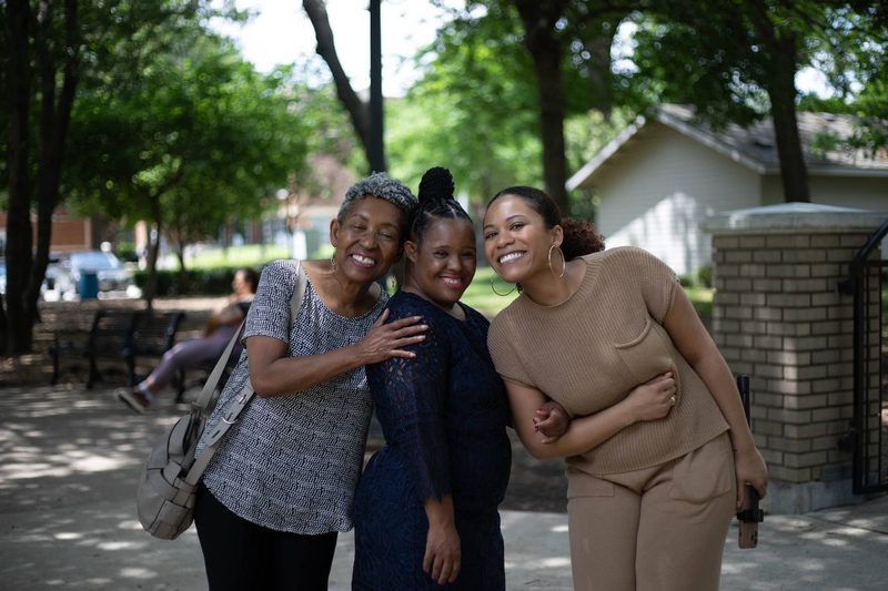 Three women laugh together on a neighborhood sidewalk.