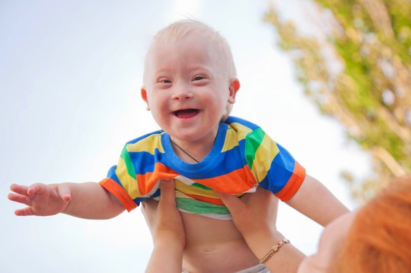 A laughing baby with Down syndrome is lifted into the air.